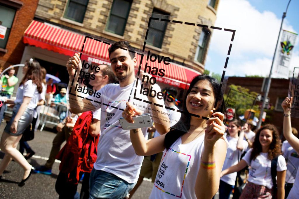 Man and woman in a diverse crowd, holding transparent squares with the "love has no labels" logos in front of their faces and wearing "love has no labels" branded t-shirts