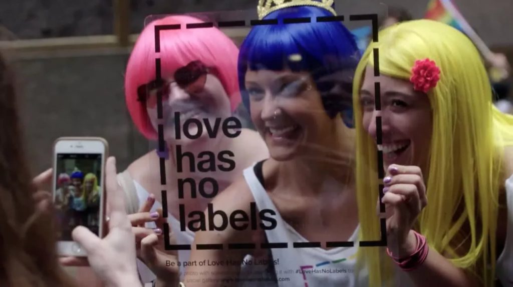 Two people with brightly colored wigs holding a transparent square with the "love has no labels" logo in front of their faces