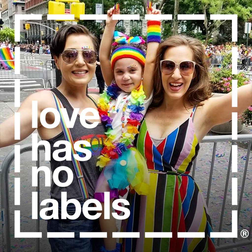 Two women with a small child, dressed in rainbow attire, with the "love has no labels" logo overlaid