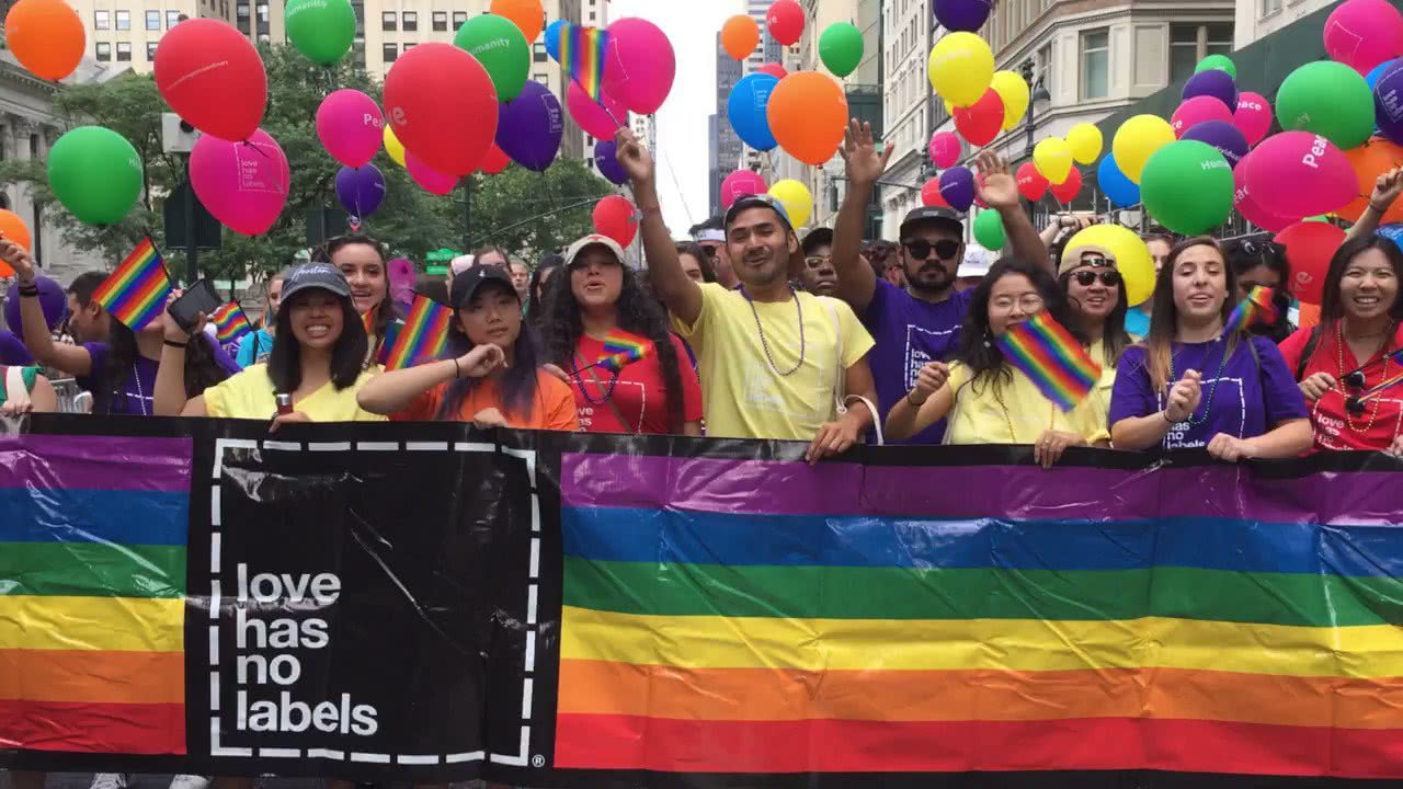 A crowd of people marching in the street with colorful balloons, dressed in colorful "love has no labels" branded t-shirts, waving rainbow flags and holding up a large rainbow banner with the "love has no labels" logo